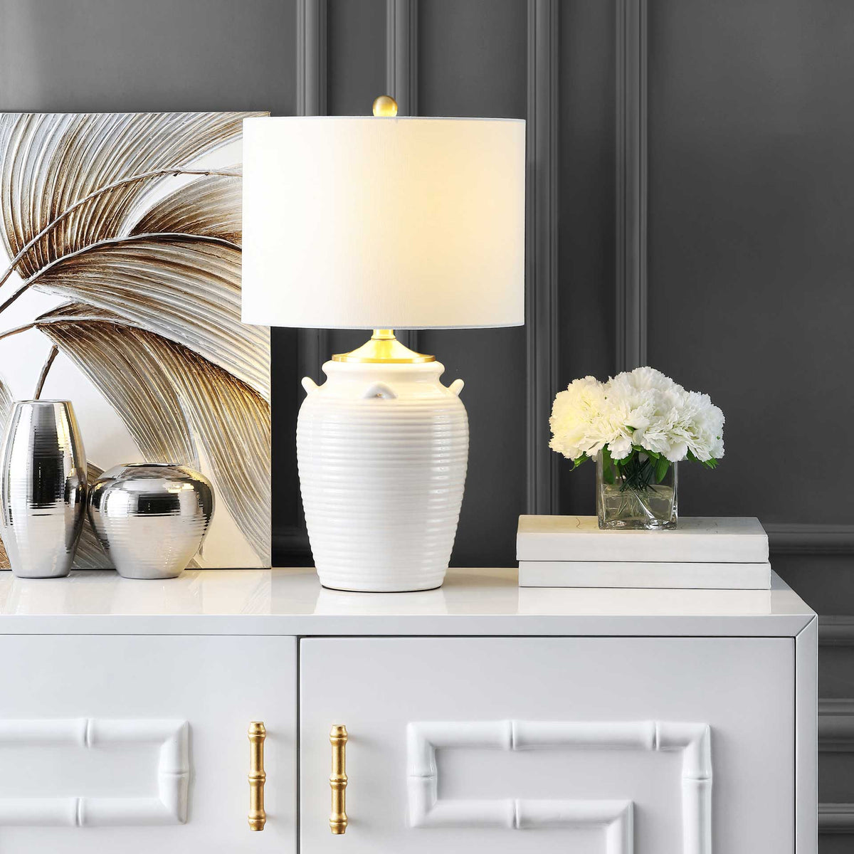 A gentle glow warms the white drum shade above the textured ivory ceramic base and brass-tone trim.