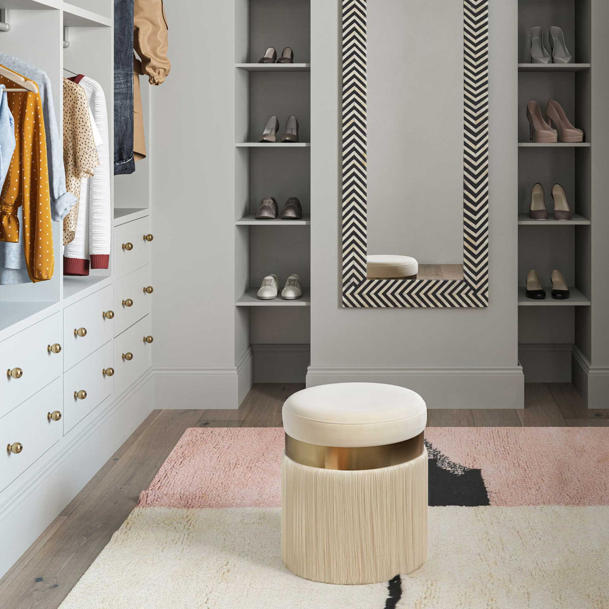 A cream velvet ottoman with long fringe offers a cozy perch in a well-appointed dressing area
