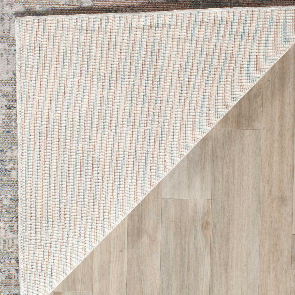 A folded corner reveals pale woven cotton backing beneath the rug's muted, multicolored surface.