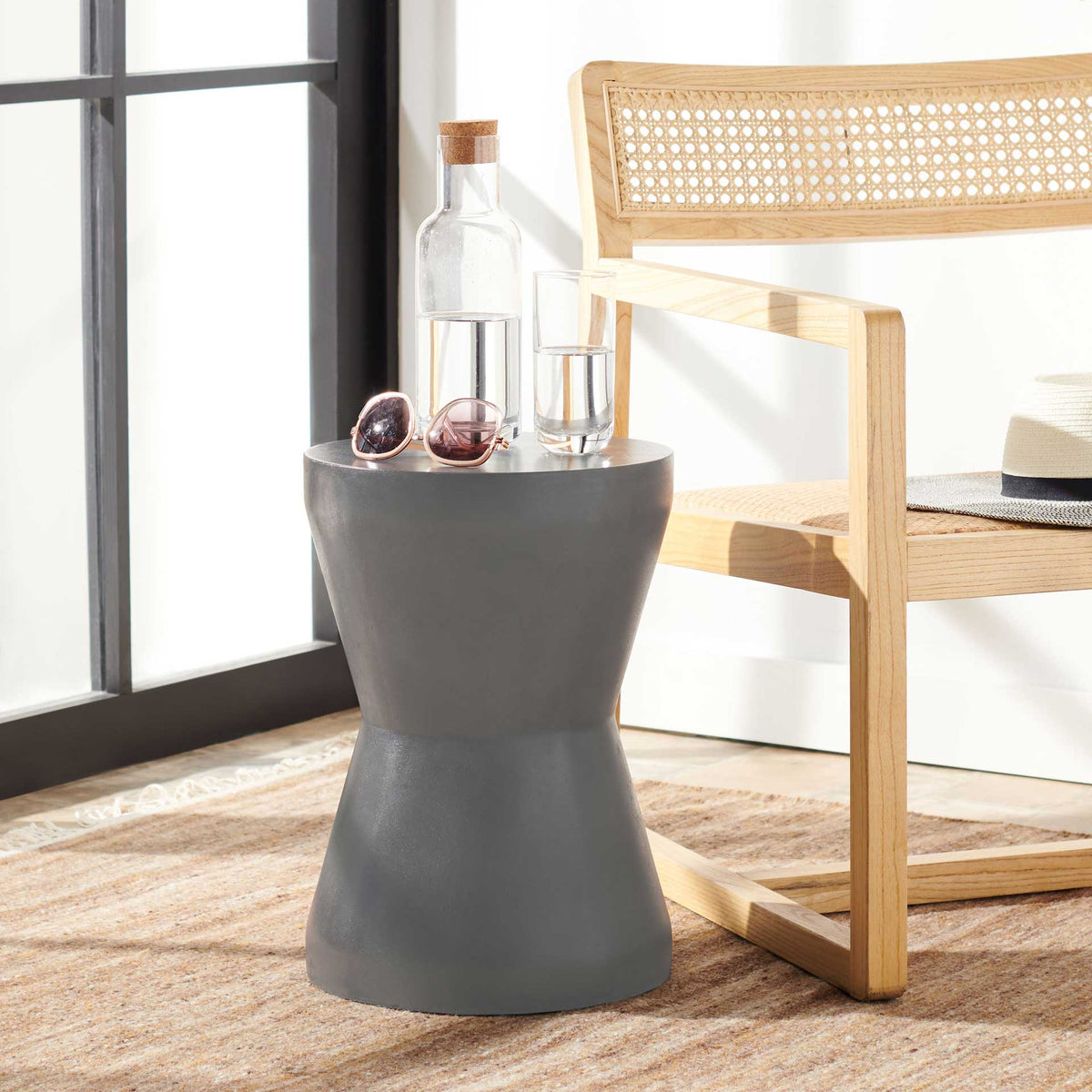 Dark gray accent table holds drinks beside a woven chair, with a slim waist and wide base